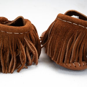 May include: A pair of brown leather moccasins with fringe. The moccasins are made of soft suede and have a simple design.
