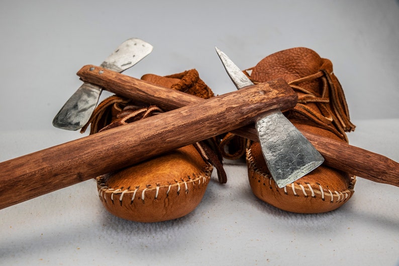 May include: Two small, brown leather moccasins with fringe and a wooden handle with two metal tomahawks attached.