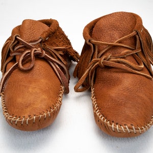 May include: A pair of brown leather moccasins with fringe and laces. The moccasins are handmade and have a traditional design.