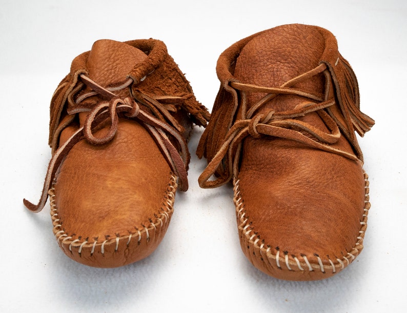 May include: A pair of brown leather moccasins with fringe and laces. The moccasins are handmade and have a traditional design.