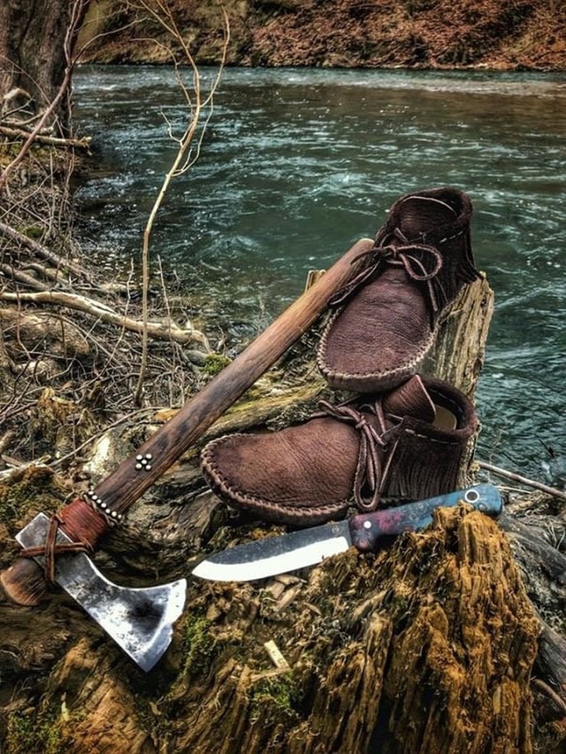 May include: A brown leather tomahawk with a wooden handle, a brown leather knife sheath, a brown leather knife, and two pairs of brown leather moccasins on a tree stump near a river.