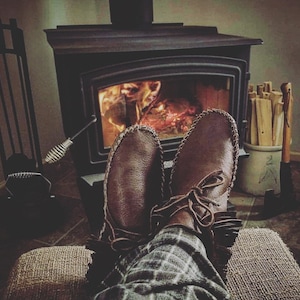 May include: A pair of brown leather moccasins with fringe detail are worn by a person sitting in front of a wood-burning stove. The stove is lit and the flames are visible. The person is wearing plaid pants.