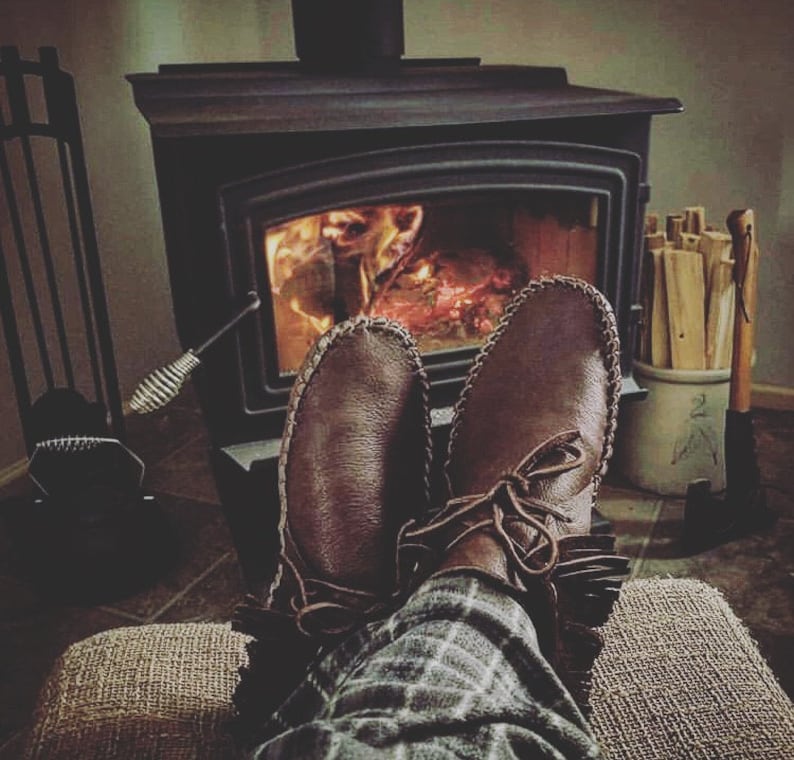 May include: A pair of brown leather moccasins with fringe detail are worn by a person sitting in front of a wood-burning stove. The stove is lit and the flames are visible. The person is wearing plaid pants.
