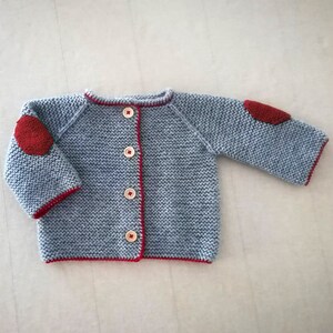 Puede incluir: Un cárdigan de punto azul claro con ribetes rojos y parches de tela roja en los codos. El cárdigan tiene cierre de botones.