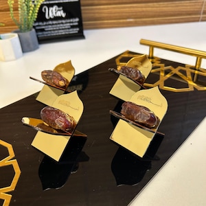 May include: Four gold-colored date holders with a mirrored finish, each holding a single date. The holders are arranged on a black, reflective surface. The holders have an arched design with Arabic script.