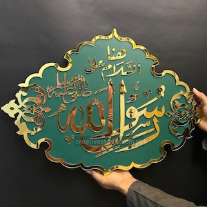 May include: A decorative wall hanging featuring gold Arabic calligraphy on a green background. The calligraphy is in a stylized script, and the piece has a gold decorative border. The wall hanging is held by a person.