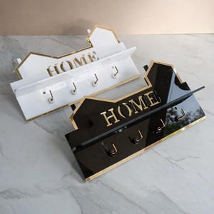 May include: Two key holders shaped like houses, one white with gold trim, the other black with gold trim. Both feature the word "HOME" in gold and have hooks for keys. A stylish home decor item.