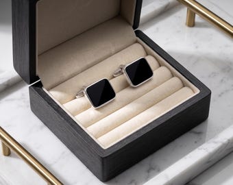 May include: A pair of square black cufflinks with silver-tone metal settings, displayed in an open black wooden box with a beige velvet interior. The box is on a white marble surface.