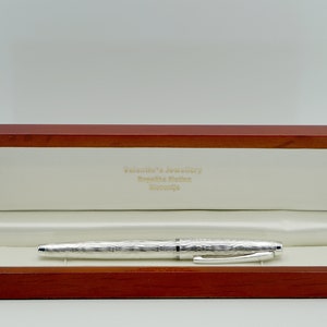May include: A silver pen with a textured finish, in a wooden box with a white interior. The box has the text "Valenti's Jewelry, Boutique Store, Greenville" on the inside.