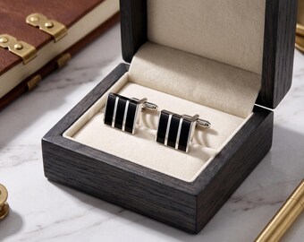 May include: A pair of square cufflinks with a black and silver striped design, presented in a dark wooden box with a cream-colored interior. The cufflinks are displayed on a marble surface, alongside a leather-bound book and a gold pen.