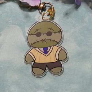 May include: A green and brown stitched character with a yellow sweater, blue tie, and black pants. The character has a stitched mouth, black eyes, and black glasses. The character is attached to a gold key chain.