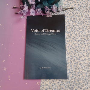 May include: A book titled "Void of Dreams" with the subtitle "Poetry and Writings Vol. 1" by Rachael Anra. The cover has a dark blue to gray gradient design. The book is on a pink and blue floral background with a floral arrangement.