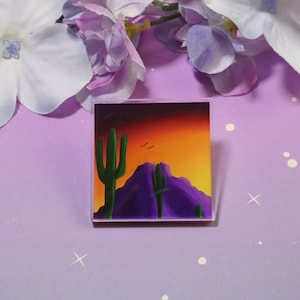 May include: A square acrylic block featuring a desert landscape painting. The artwork depicts a purple mountain range, green cacti, and a vibrant sunset with orange, yellow, and red hues. Two birds are flying in the sky.