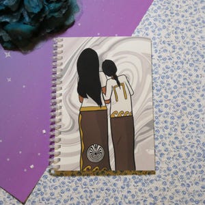 May include: Spiral-bound notebook featuring an illustration of two figures with long hair, standing together. They are wearing white tops with gold accents and brown skirts. The background has a swirling gray and white pattern.