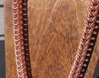 Puede incluir: Un collar de cadena de oro rosa hecho de anillos de metal entrelazados. El collar se muestra en un soporte de madera.