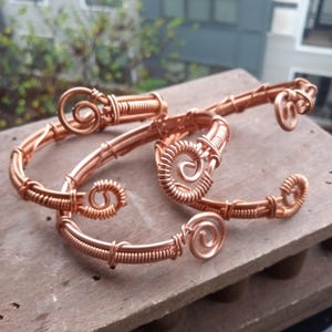 Puede incluir: Tres pulseras de alambre de cobre con diseños en espiral. Las pulseras están apiladas una encima de la otra sobre una superficie de madera.