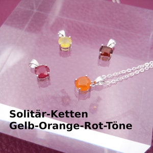 Puede incluir: Cuatro colgantes de plata con piedras preciosas de diferentes colores: amarillo, naranja, rojo y marrón. Los colgantes se muestran sobre una superficie transparente con un collar de cadena de plata. El texto "Solitär-Ketten Gelb-Orange-Rot-Töne" es visible debajo de los colgantes.