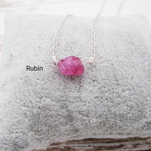 Könnte beinhalten: Halskette mit silberner Kette und einem Anhänger aus rohem, leuchtend rosa Rubin. Das Wort "Rubin" ist in schwarzer Schrift gedruckt. Die Kette liegt auf einer weichen, grauen Oberfläche.
