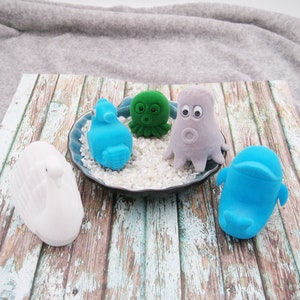 May include: Five small plush toys in the shape of sea creatures. A white swan, a blue seahorse, a green octopus, a gray octopus, and a blue whale. The toys are sitting on a white and gray decorative plate.