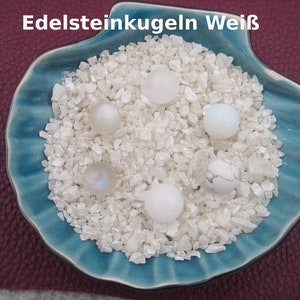 Könnte beinhalten: Eine blaue Keramik-Muschelschale gefüllt mit weißen Edelsteinsplittern und sechs weißen Edelsteinkugeln. Der Text "Edelsteinkugeln Weiß" ist in weiß auf weißem Hintergrund über der Schale geschrieben.