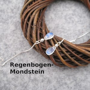 Könnte beinhalten: Ein Paar silberne Ohrhänger mit regenbogenfarbenen Mondstein-Tropfen. Die Ohrringe liegen auf einem braunen geflochtenen Untergrund. Der Text "Regenbogen-Mondstein" ist unterhalb der Ohrringe sichtbar.