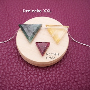 Op de afbeelding: Drie driehoekige edelsteen hangers in verschillende maten. De grootste hanger is groen, de middelste hanger is geel en de kleinste hanger is roze. De hangers bevinden zich op een houten display met de tekst "Dreiecke XXL" en "Normale Größe".
