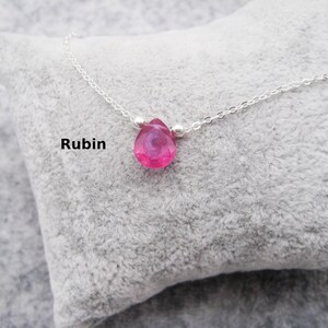 Pode incluir: Um colar de corrente prateada com um pingente de pedra preciosa rosa brilhante, facetada e em forma de gota. A palavra "Rubin" está impressa em preto à esquerda. O colar é exibido em uma superfície cinza e macia.