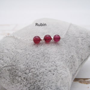 May include: A silver chain necklace with three red ruby beads. The necklace is on a grey, fuzzy surface. The word "Rubin" is written above the necklace.
