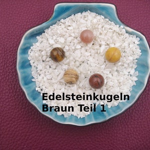 Könnte beinhalten: Fünf Edelsteinkugeln in einer blauen Muschelformschale auf einem burgunderfarbenen Untergrund. Die Kugeln sind braunes Tigerauge, rosa, braunes Holz, rot und gelb. Der Text "Edelsteinkugeln Braun Teil 1" ist am Boden der Schale sichtbar.