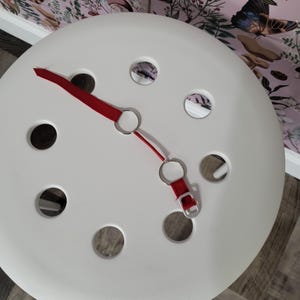 K&ouml;nnte beinhalten: Ein wei&szlig;er Hocker mit einem roten Riemen und silbernen Ringen. Der Hocker hat mehrere runde L&ouml;cher in der Oberseite.