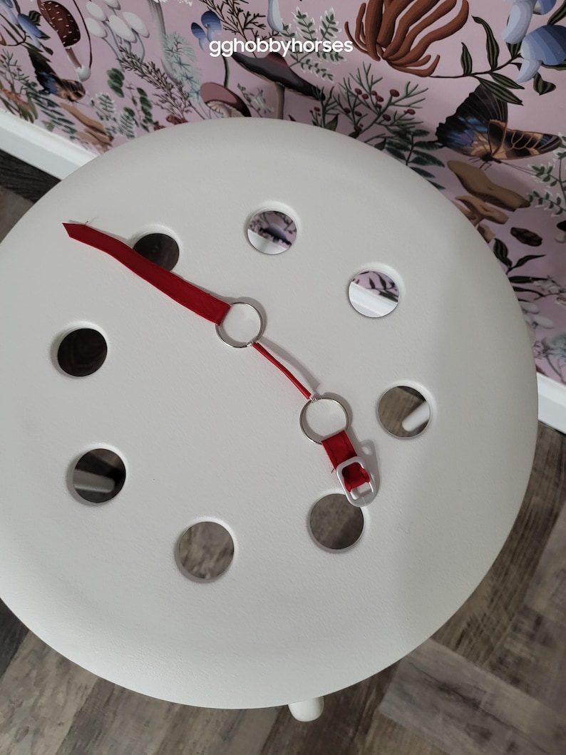K&ouml;nnte beinhalten: Ein wei&szlig;er Hocker mit einem roten Riemen und silbernen Ringen. Der Hocker hat mehrere runde L&ouml;cher in der Oberseite.