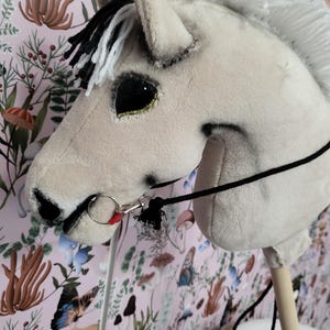 K&ouml;nnte beinhalten: Ein wei&szlig;es und graues Pl&uuml;sch-Steckenpferd mit schwarzer M&auml;hne und Schweif. Das Pferd hat ein schwarzes Halfter mit einem roten und silbernen Druckknopf. Das Pferd steht auf einem Holzstab.
