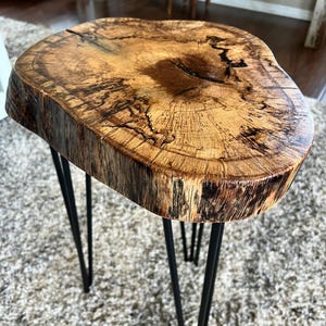 May include: A handcrafted side table with a natural wood top and black hairpin legs. The wood top has a unique, organic shape and a rich, warm brown color with visible grain patterns. The table is approximately 18 inches in diameter and 18 inches tall.