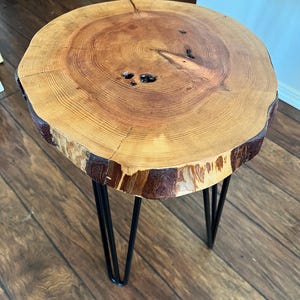 May include: A round wooden side table with a natural wood grain top and black hairpin legs. The tabletop is approximately 18 inches in diameter and features visible growth rings and knots, showcasing the wood's natural texture and color variations.