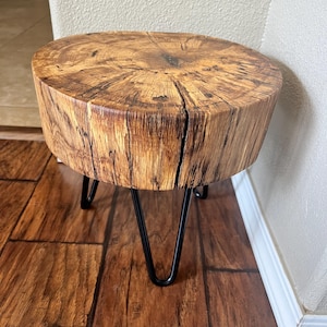 May include: A round wooden table with a natural wood finish and black hairpin legs. The table is made from a tree trunk slice and has a rustic look.
