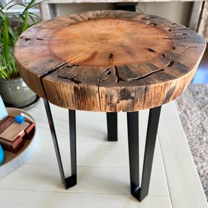 May include: A round wooden table with a natural wood finish and black metal legs. The table top has a unique, rustic look with knots and grain patterns.