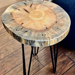 May include: A round wooden side table with a natural wood finish and black hairpin legs. The table top is a slice of a tree trunk, showing the natural grain and rings.