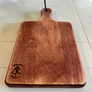 May include: A rectangular wooden cutting board with a handle and a dark brown finish. The board has a small, dark logo in the lower left corner and a dark green loop for hanging. The wood grain is visible.