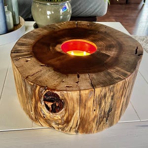 May include: A wooden candle holder made from a tree trunk with a circular opening in the center for a candle. The candle holder is a natural wood color with a dark brown stain. The candle holder is sitting on a white table.