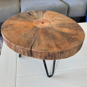May include: A round wooden table top with a natural wood grain pattern and a dark brown stain. The table top is supported by three black metal hairpin legs.