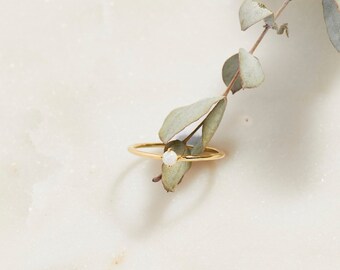 Puede incluir: Un delicado anillo de oro con una pequeña gema blanca y redonda. El anillo se encuentra sobre una superficie de mármol blanco, con una ramita de vegetación. El diseño sencillo del anillo lo convierte en una joya versátil.