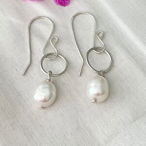 Op de afbeelding: Een paar zilveren oorhangers met grote, onregelmatige witte parels. De oorbellen hebben een eenvoudig ontwerp met een haak en een cirkelvormig element dat verbonden is met de parel. De parels hebben een glanzend, glad oppervlak.