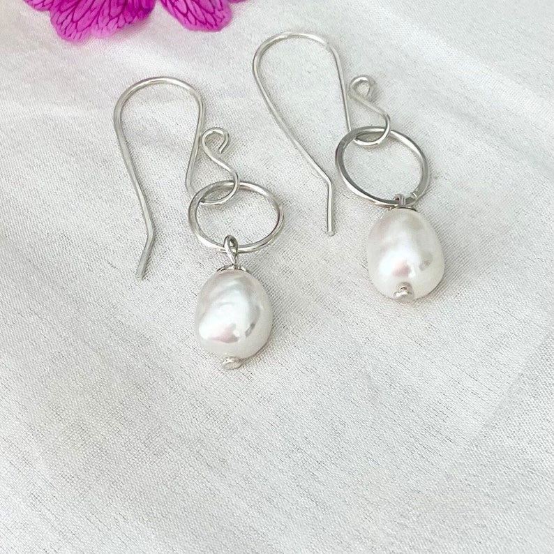Op de afbeelding: Een paar zilverkleurige oorhangers. Elke oorbel heeft een grote, witte, peervormige parel die aan een kleine zilveren ring hangt. De ring is bevestigd aan een gebogen zilveren haak. De oorbellen worden op een wit oppervlak getoond.