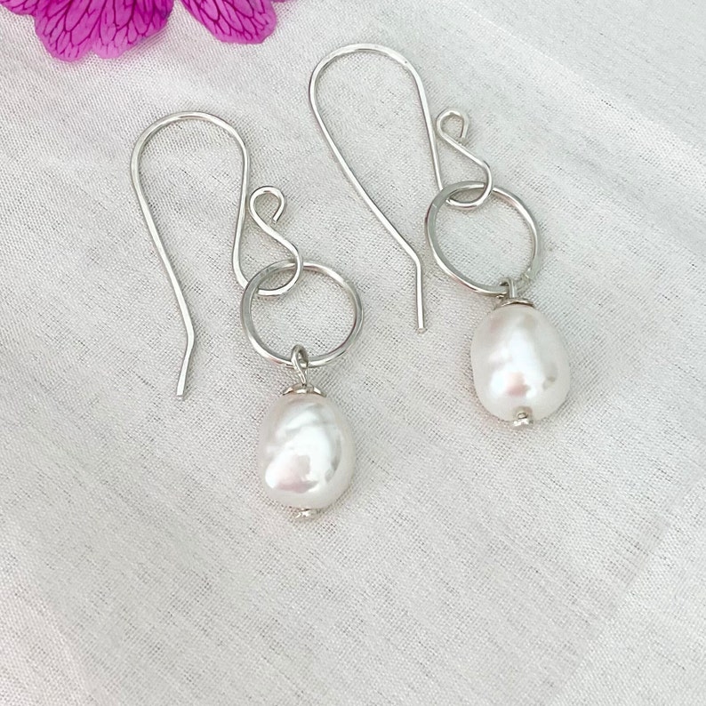 Op de afbeelding: Zilveren oorhangers met grote, onregelmatige witte parels. De oorbellen hebben een eenvoudig ontwerp met een haak en een kleine ring die verbinding maakt met de parel. De achtergrond is een witte, getextureerde stof, met een roze bloem in de linkerbovenhoek.