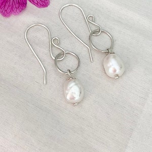 Op de afbeelding: Een paar zilverkleurige oorhangers met een grote, witte barokparel die aan een kleine zilveren ring hangt. De oorbellen hebben een eenvoudig haakontwerp. Op een witte achtergrond met een paarse bloem.