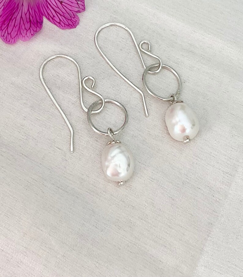 Op de afbeelding: Een paar zilverkleurige oorhangers met een grote, witte barokparel die aan een kleine zilveren ring hangt. De oorbellen hebben een eenvoudig haakontwerp. Op een witte achtergrond met een paarse bloem.