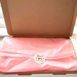 Op de afbeelding: Een open bruine kartonnen doos met roze vloeipapier. Een ronde sticker met de tekst "thank you" en "for supporting my small business" is op het papier geplakt. De doos staat op een wit oppervlak.