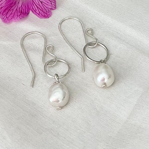 Op de afbeelding: Een paar zilveren oorhangers. Elke oorbel heeft een grote, onregelmatige witte parel die aan een kleine zilveren ring hangt. De oorbellen hebben een eenvoudig ontwerp met een gebogen haak.
