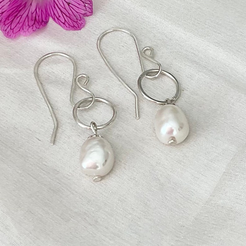 Op de afbeelding: Een paar zilveren oorhangers. Elke oorbel heeft een grote, onregelmatige witte parel die aan een kleine zilveren ring hangt. De oorbellen hebben een eenvoudig ontwerp met een gebogen haak.