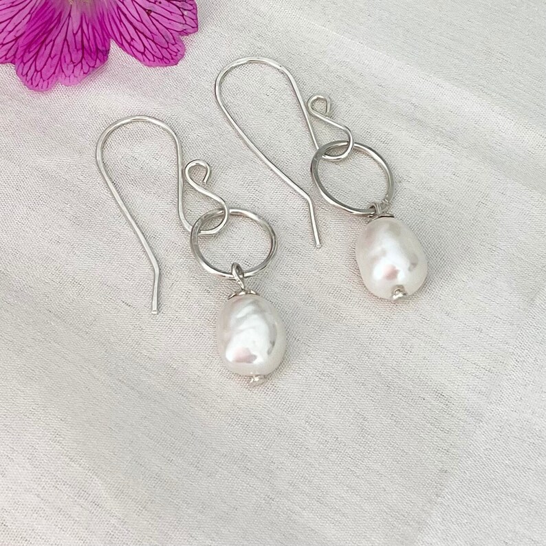 Op de afbeelding: Een paar zilveren oorbellen met grote, witte, druppelvormige parels. De oorbellen hebben een eenvoudig ontwerp met een haak en een kleine ring die met de parel verbonden is. Op de achtergrond is een roze bloem te zien.